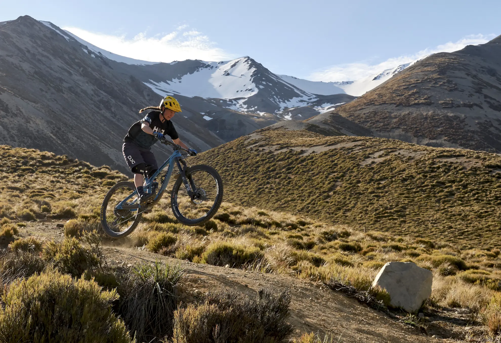 Female mountain biking jumping as she rides down a trail with snowy mountains in the background
