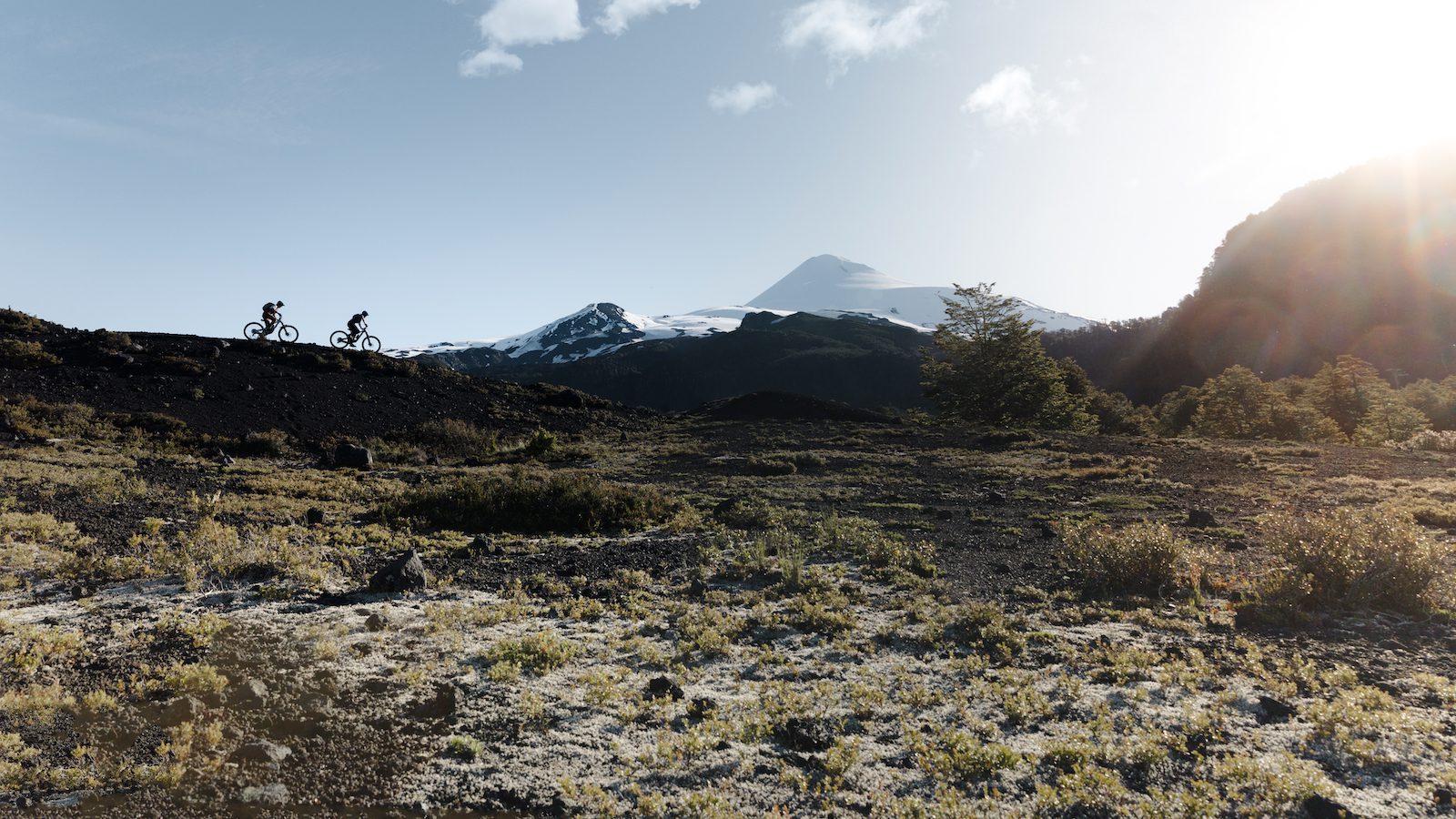 Two riders are silhouetted as they ride along a ridgeline with snowcapped mountains in the background