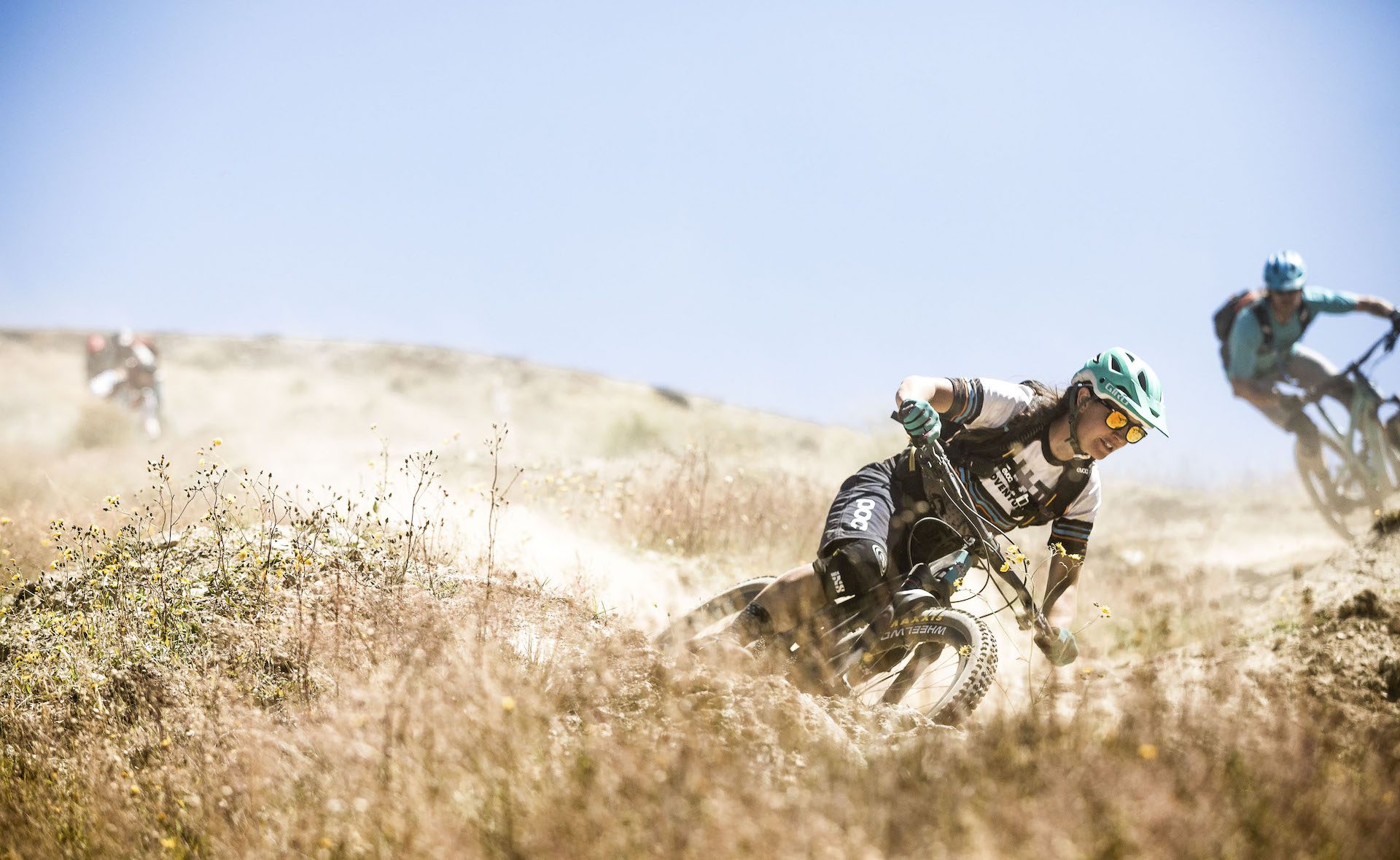 Two mountain bikers riding down a dusty trail, with blue sky and long grass in the foreground